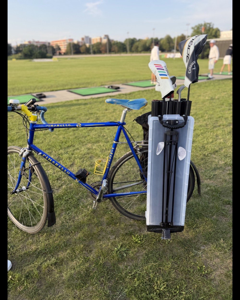 The VeloCaddie prototype clipped onto the rear rack of a blue Pinarello road bike, parked on the grass at a driving range, clubs and headcovers standing out of the top.