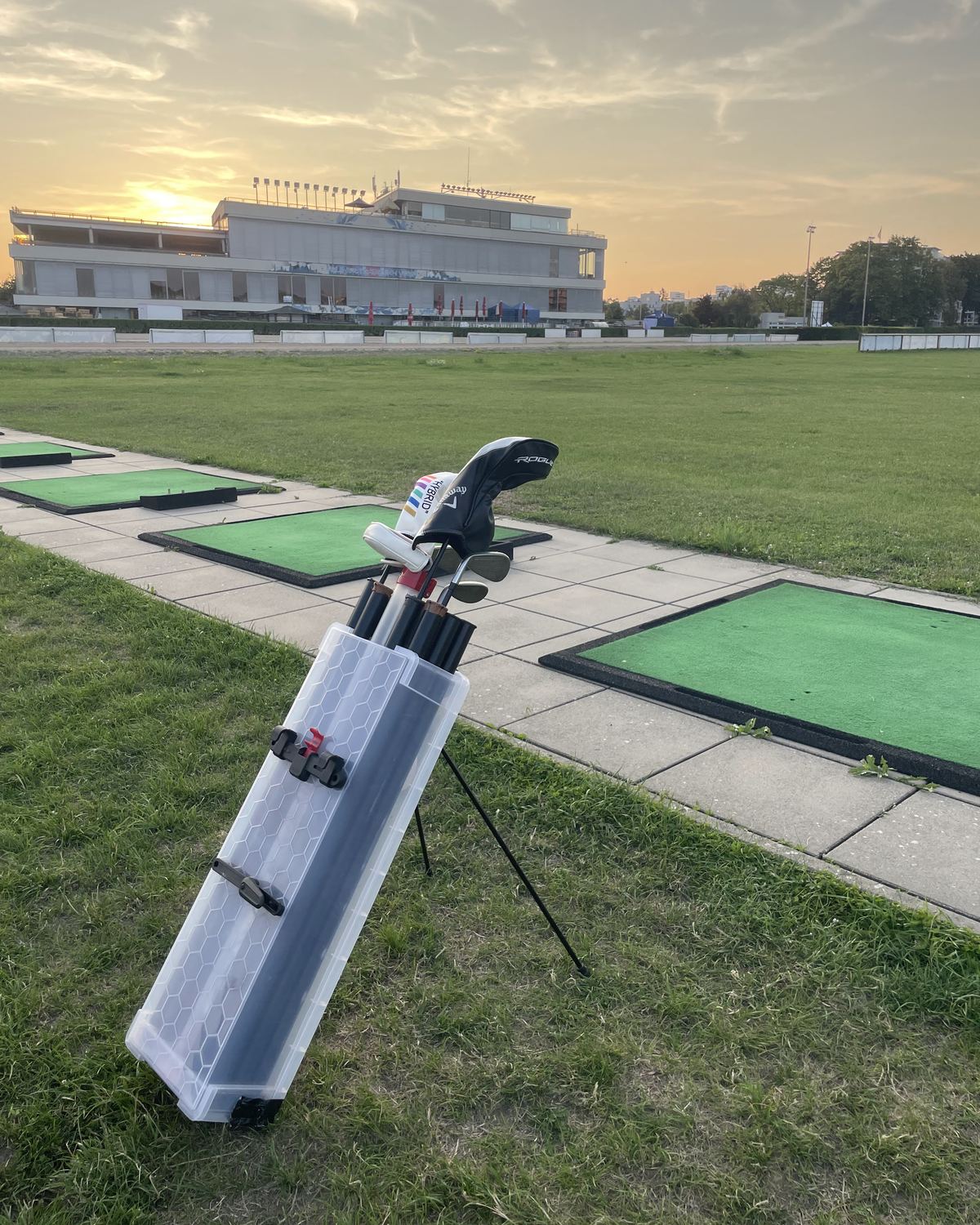 The VeloCaddie v3 prototype standing on its fold-out leg beside the practice mats of a driving range, clubs in it, the clubhouse and a low evening sun behind.