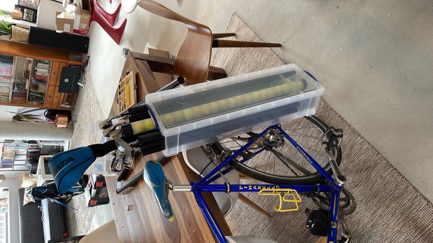 A clear plastic storage box holding golf clubs, standing next to a blue road bike indoors.
