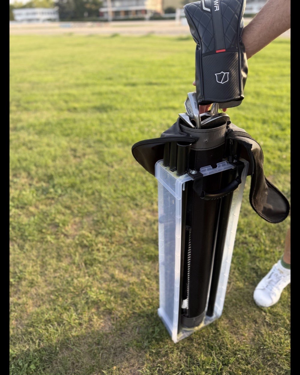 The case lifted by hand: a black soft golf bag with its rain hood sits inside the clear case, club tubes visible beside it.