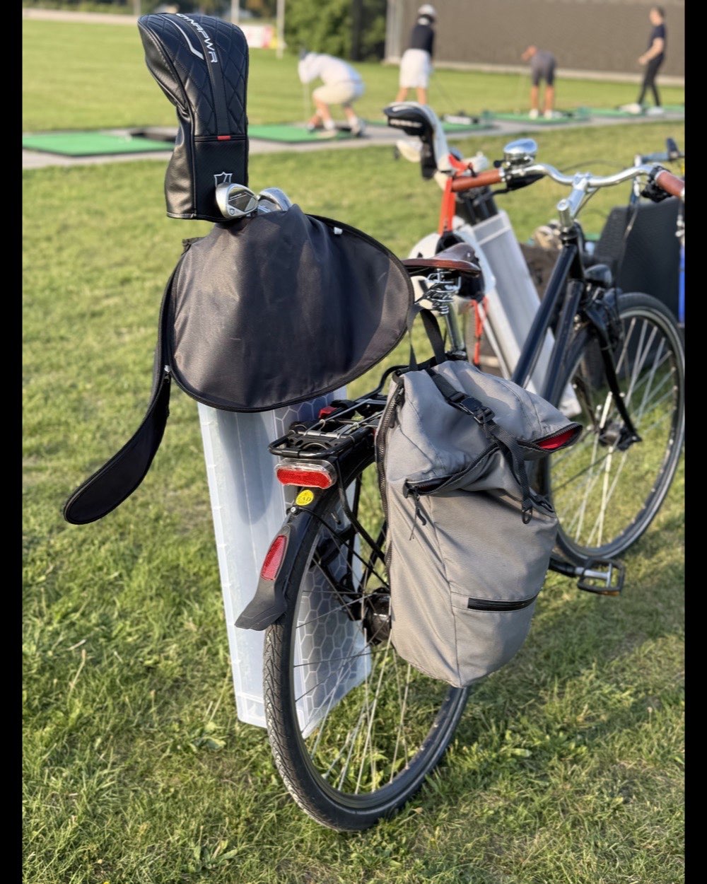 Rear view of the combination mounted on a city bike's rear rack next to a grey pannier, the black rain hood covering the clubs.