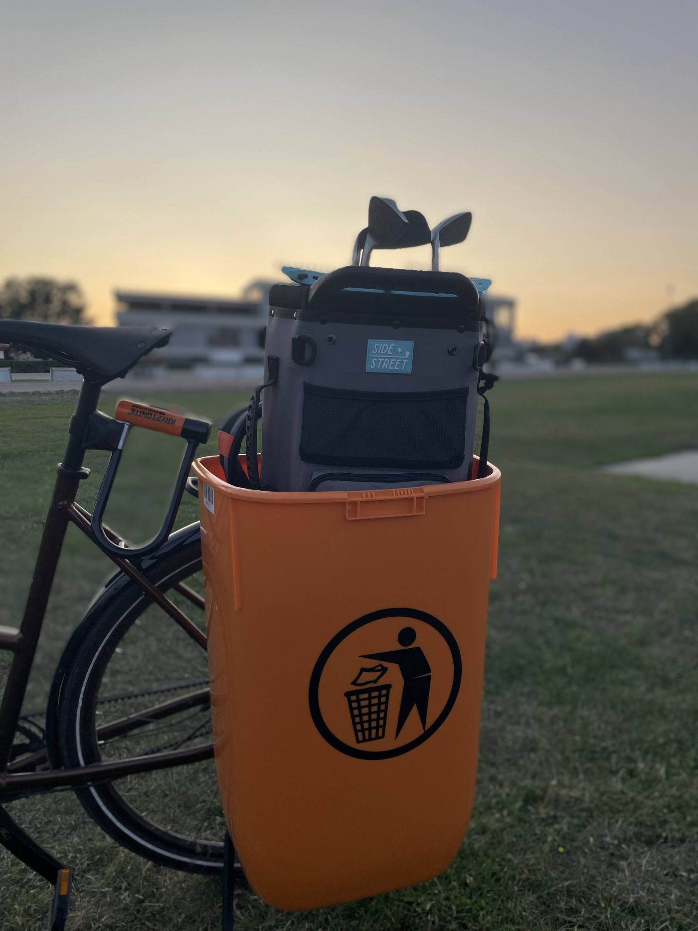 The first Dockingbox shell on the rear rack of a bicycle at a driving range at sunset: an orange rigid shell with a grey golf bag and clubs docked inside.